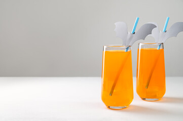 Halloween Mocktails. Two glasses of orange non-alcoholic cocktail with ice for Halloween party on a white table. Straws are decorated with cut paper bats. Selective focus, copy space.