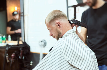 Obraz premium Side view of a man in a stripe cape visiting a barber shop, getting a trendy hairstyle, Unrecognizable hairdresser is mirrored on the blurred background