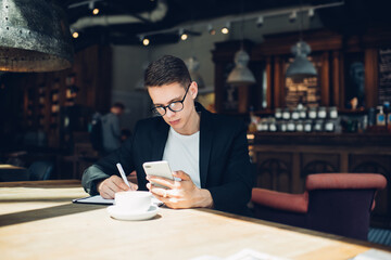 Pondering student with smartphone in hand studying textbook while sitting at cafe