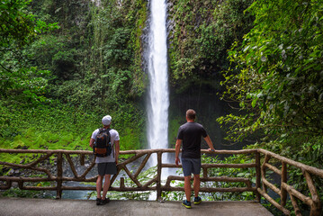 Two tourists looking at the La Fortuna Waterfall in Costa Rica - obrazy, fototapety, plakaty