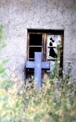 cross of a tombstone leans at a broken window