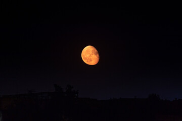 Bright orange moon in waning gibbous phase rising up on dark clear night sky above dark city...
