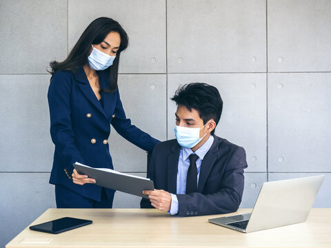 Asian business man and woman wearing suit and protective face masks using computer on desk, meeting and working together in office.