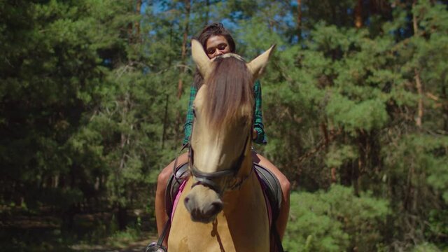 Portrait of charming positive african female in checkered shirt and shorts sitting in saddle on brown purebred horse, petting gently with love and care neck of horse while riding horseback in wood.