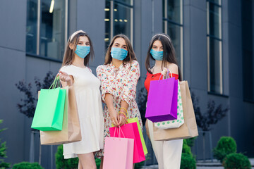 Women in casual dresses, top and pants wearing masks to protect coronavirus pandemic standing in front of the shopping mall with colored bags in hands.