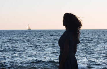 Fototapeta premium Silhouette of a woman at the beach looking across a blue ocean to a sailboat on the horizon.