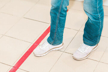 Person stand in red line, legs close-up. Attention line on the floor of the store to maintain social distance. Concept of the coronavirus pandemic and prevention measures