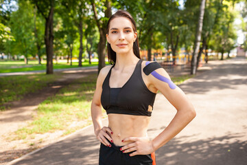Fototapeta premium Portrait of attractive muscular brunette woman wearing black sports outfit, looking at camera. Young smiling female athlete posing with hands on waist, colorful kinesiotaping on body.