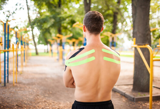 Unrecognizable young caucasian professional bodybuilder with black and green kinesiological tapes on naked muscular shoulders posing at sports ground.