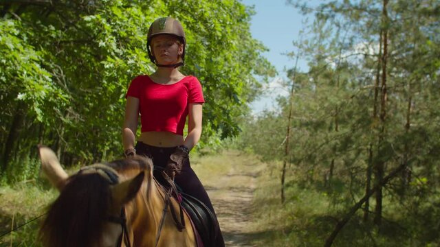 Portrait of concentrated young female rider in helmet with whip riding horseback on forest trail. Girl jockey in horse riding equipment and riding clothes enjoying horseback ride in summer nature.