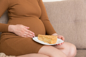 Close up of hungry pregnant woman is eating a piece of tasty cake relaxing on the sofa at home. Sweet cravings during pregnancy
