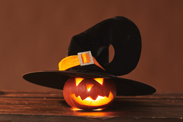 Close-up studio shot of cute Jack O' Lantern wearing black witch hat with candle glowing inside of it, brown background