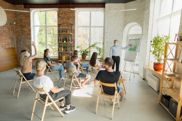 Male speaker giving presentation in hall at university workshop. Audience or conference hall. Young students, participants in audience wearing face mask. Scientific conference event, training