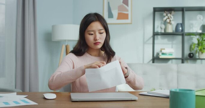 Charming Asian girl recieving response from university, college, job. Excited young woman opening and reading letter, getting happy and rejoicing at home. Education, admission, occupation.
