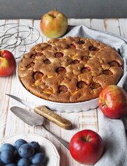 Freshly baked summer round pie with apples and plums in a white ceramic form on a gray background.Food background.