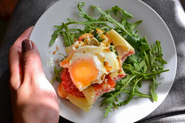 Tasty breakfast. Toast with egg, cheese and sausage. Sandwich in a white plate with arugula. View from above. Light background. Closeup. Woman holding plate with breakfast