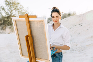 Young woman artist painting on canvas on a easel, holding a palette.