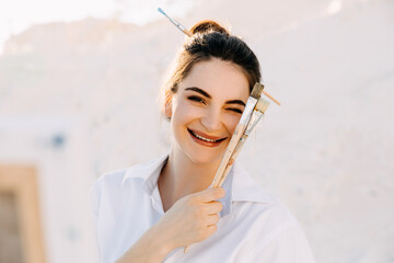 Young woman artist posing with paintbrushes, smiling and winking.