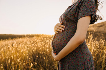 Closeup of a pregnant woman in a field with dry grass, holding hands on her belly.