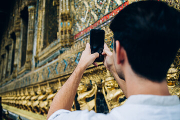 Back view of man holding mobile phone shooting video on mobile phone visiting golden asian temple, male traveler using smartphone camera taking photo for share in travelblog during journey.