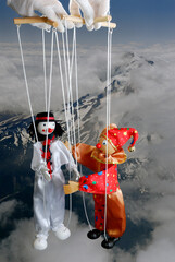 Clown Marionettes about to embrace and white gloved pupeteer with a snow covered mountain background