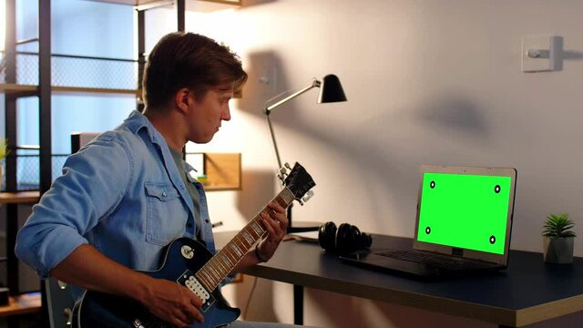 leisure, music and people concept - young man or musician with green chroma key screen on laptop computer playing bass guitar sitting at table at home