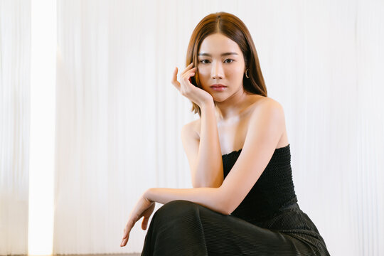 Portrait of a stunning fashionable beautiful sexy Asian woman in stylish strapless dress clothes with hand on chin and looking at camera. Fashion and beauty concept. Lady slim wears black fit clothes.