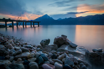 A tranquil pier or a bridge on lake of Garda with stones in the foreground and sunset behind rich big mountains. Concept of calmness on a lake.