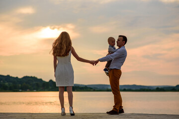 Father mother and their one year old son. The father holds his son with one hand and his wife with the other.