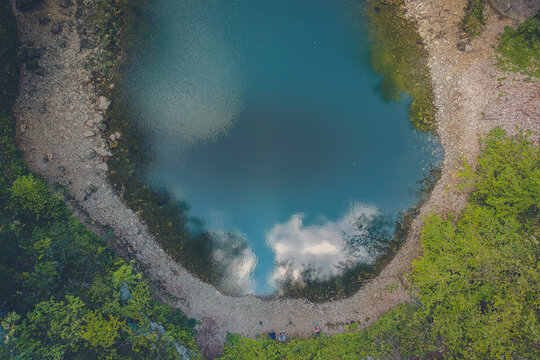 A round small lake called Divje jezero in Slovenia, surrounded with pebbles and still water on a sunny day with blue skies, hiding between thick green forest.