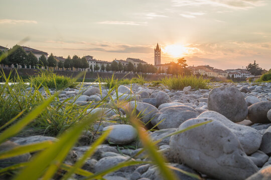 View of Basilica di San Zeno Maggiore in Verona in sunset looking above the Adige river with stones and grass on the river bank. Romantic picuture of a church in verona.