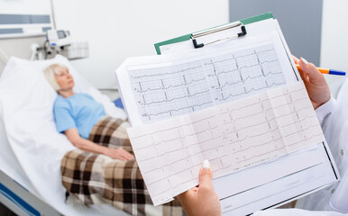 Experienced doctor holding electrocardiogram results of an elderly woman. Senior patient lies in a...