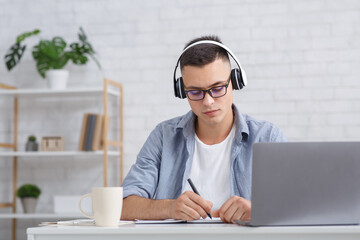 Busy young man with glasses makes notes near laptop in living room interior