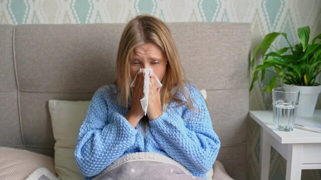 Upset young woman suffering from influenza at home. Female blows her nose holding tissue handkerchief, having symptoms of chronic sinusitis disease, seasonal allergy or cold or flu.