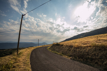 Panorama of a beautiful summer landscape with sun, clouds and yellow fields in France. Rural scenic asphalt road in a curve visible going around the corner