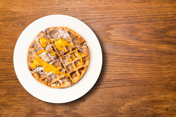 Tasty waffles sprinkled with powdered sugar and apricot jam, lying on a plate, on a wooden table. View from above
