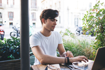 Brunette young male student typing on laptop computer making research for education online course, skilled caucasian man freelancer working on project remotely sitting on cafe terrace with technology