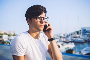 Serious handsome 20s male in spectacles making smartphone call sitting in city bay with yacht concentrated on conversation, pensive man traveler using roaming connection for talking on cellular