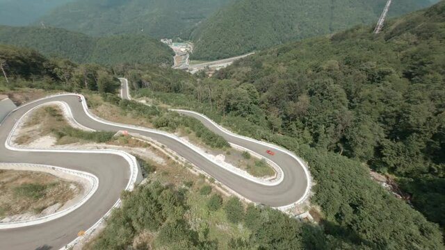 Winding road in summer highlands from sport FPV drone. Convertible car driving along curved serpentine road in Sochi, Krasnaya Polyana. Travel in mountain, covered summer forest aerial cinematic view