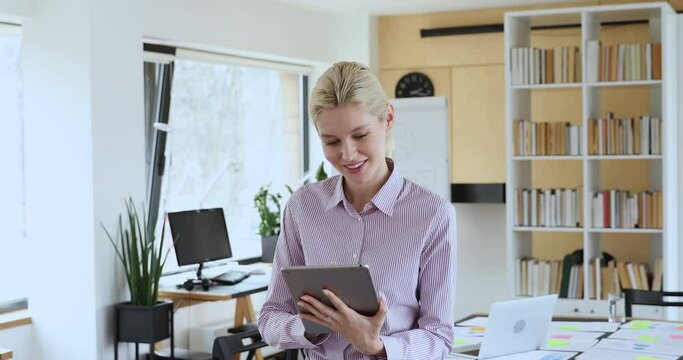 Attractive woman working using tablet standing in modern office room. Make presentation on device smiling feel satisfied by work done, having distance remote communication with company clients concept