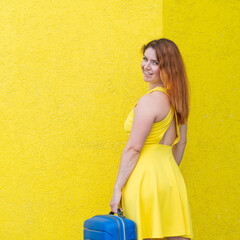 A woman in a yellow dress turns around and holds a blue suitcase on color background