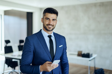 Smiling businessman texting on cell phone in the office and looking at camera.