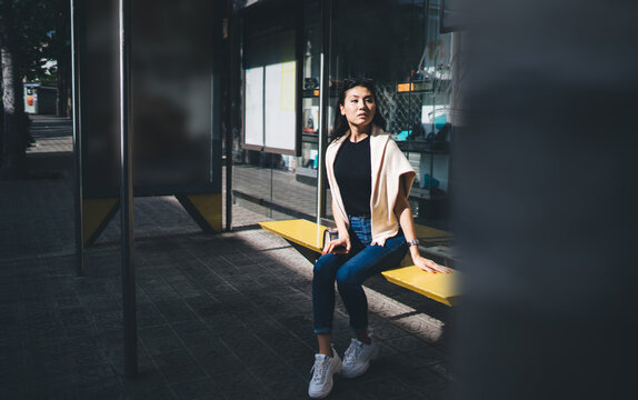 Young asian female in casual wear sitting on bench waiting for transport in evening in megalopolis, pensive hipster girl looking for bus or tram on station due to timeline schedule for transporting