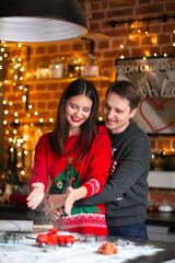 Young brunette asian woman and white man in Christmas sweaters making cookies together on the kitchen. Lights background.