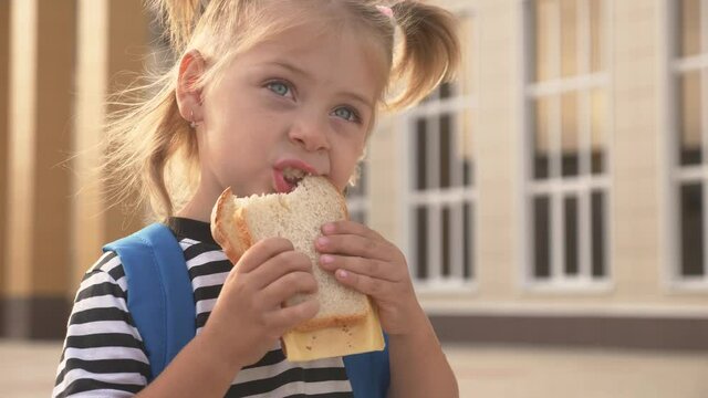 Happy kid eating a sandwich outdoors at school. Healthy school breakfast for the child. Hungry kid eating outside on the background of the school. Street fast food. Food for lunch. A delicious burger