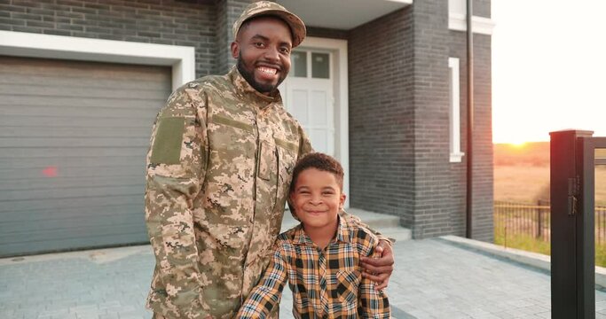 Portrait of handsome happy African American man with small cute boy hugging and smiling to camera outdoor at house. Father soldier coming back home from army and posing to camera with little son.