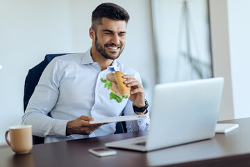 Happy businessman eating sandwich and using laptop on a break in the office.