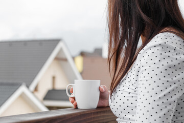 Young woman in polka dot T-shirt with a white cup of tea or coffee standing on the balcony. Morning...