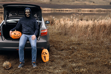 An unrecognizable adult man wearing a skull mask sits in the trunk of a car with carved pumpkins for Halloween, outdoors. Trick or trunk. Copy space.