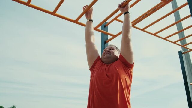 Obese person doing exercises in city park, fat man hanging on the street trainer slowmo